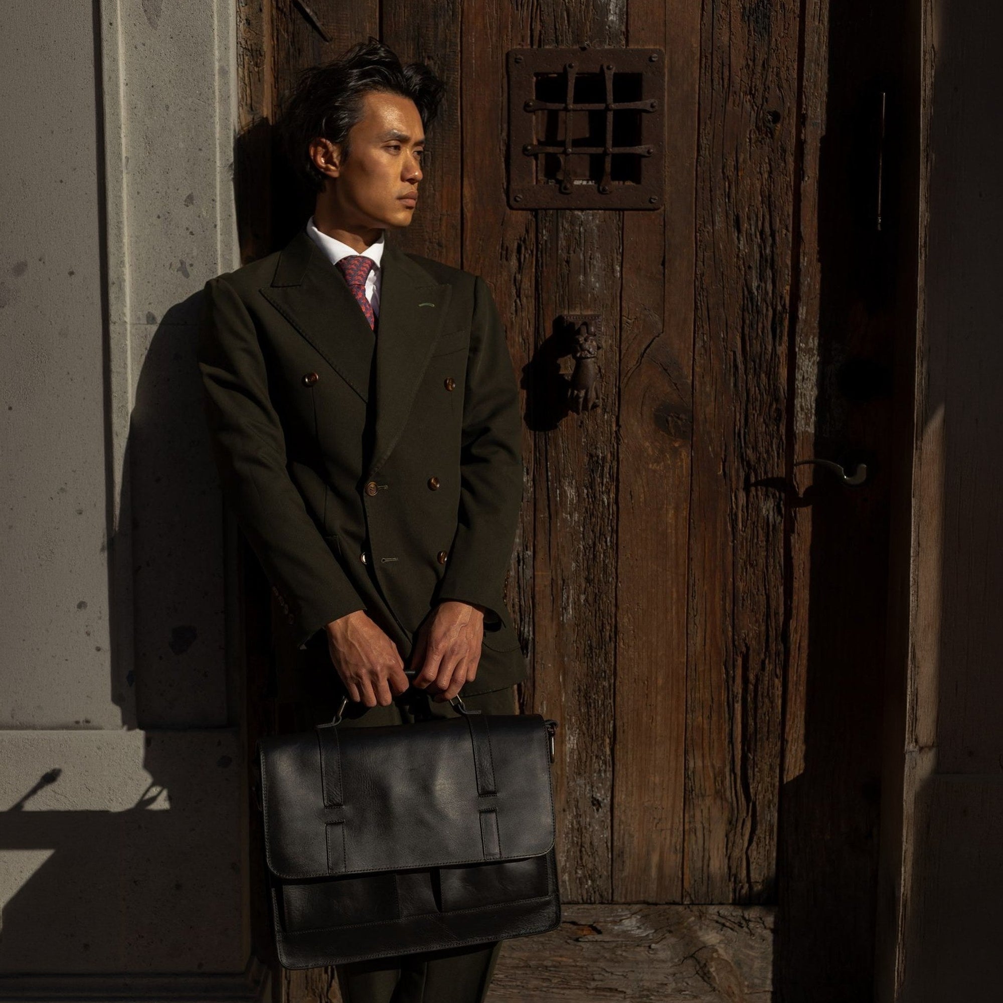 Man in a dark suit holding the Jefferson Leather Messenger Bag in Vintage Black at a wooden door.