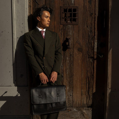 Man in a dark suit holding the Jefferson Leather Messenger Bag in Vintage Black at a wooden door.