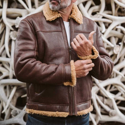 Bearded man sporting a brown shearling leather jacket, posing with confidence against an antler backdrop.