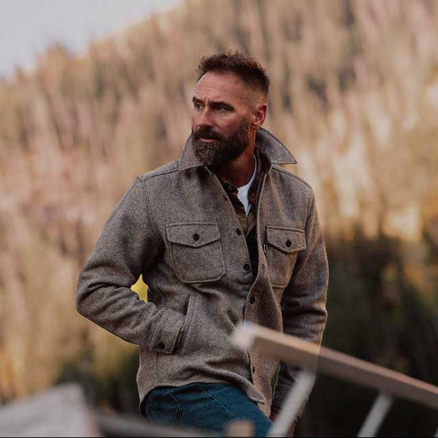 Brown wool Shirt jacket,  textured brown overshirt and blue jeans, standing outdoors in front of a soft-focus background of dry grass and trees.