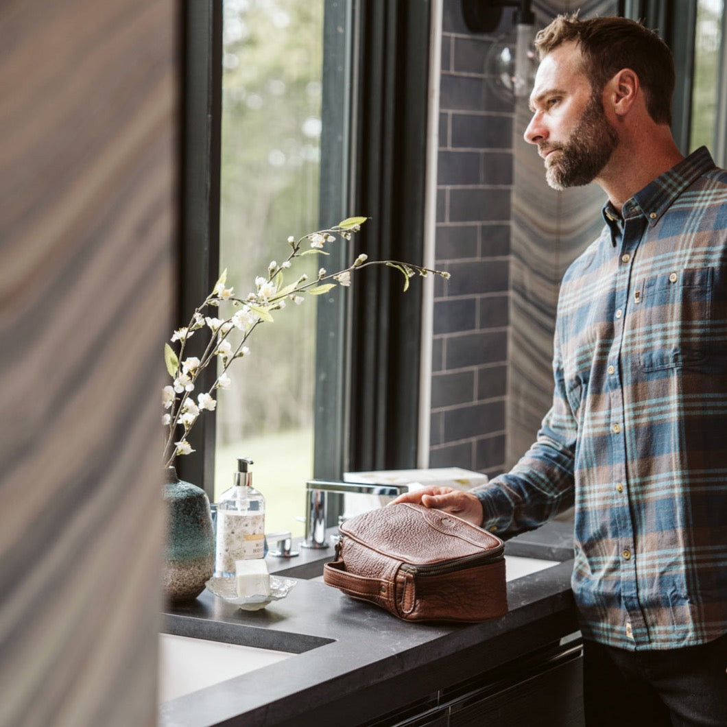 Man standing by a bathroom window, holding a brown leather dopp kit, partially open to display neatly arranged toiletries