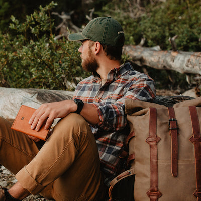 Man sitting outdoors with a backpack and leather-bound book, surrounded by nature.