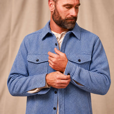A man wearing a textured blue herringbone overshirt over a cream shirt, adjusting the front buttons. The background is a soft beige fabric backdrop.