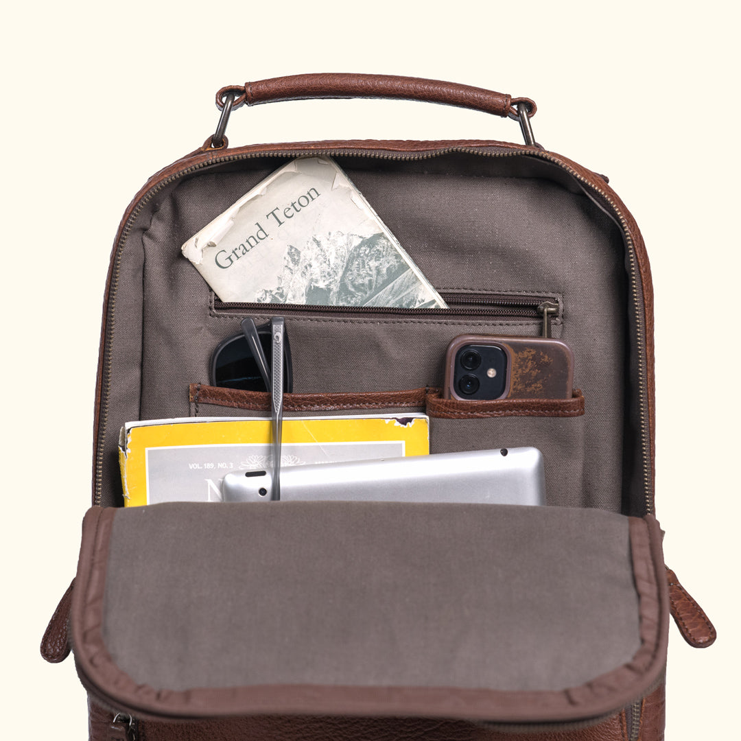 Organized brown leather backpack interior packed with a laptop, smartphone, pen, notebook, and vintage postcard for travel-ready efficiency.