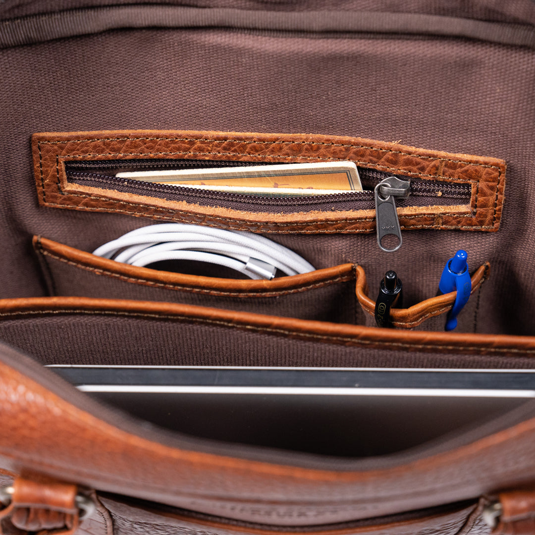 Interior view of a leather briefcase with organized compartments for pens, cables, and a laptop.