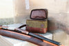 Waxed canvas hunting bag with leather accents, open beside a shotgun on a metal background.