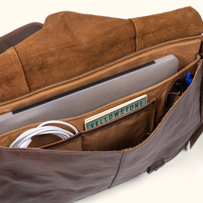 Interior view of a brown leather messenger bag, showcasing organized compartments for laptop, cables, and pens.