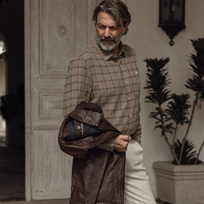 Man in the Windsor Cotton Shirt in Timber Pane carrying a leather jacket through a courtyard
