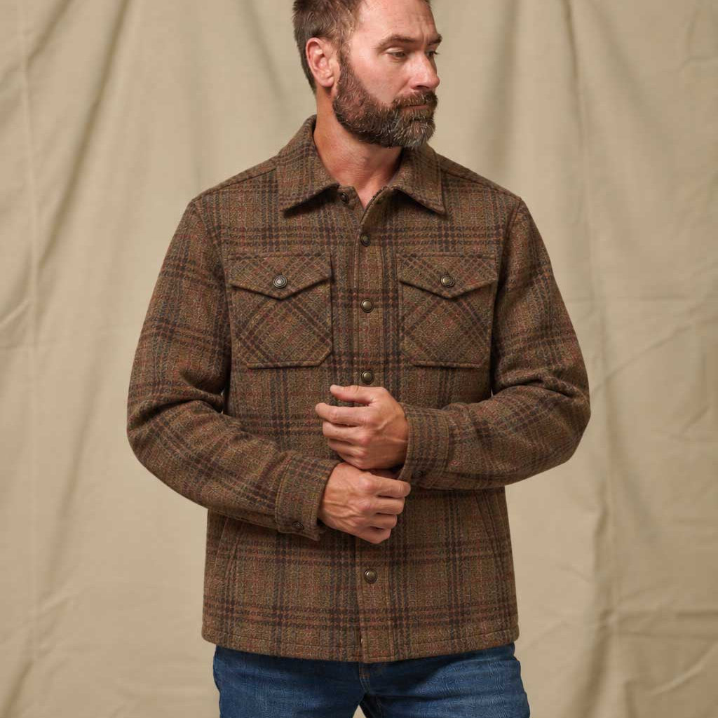 A man with a beard wearing a brown and black plaid button-up flannel shirt jacket and blue jeans, standing against a beige fabric backdrop.
