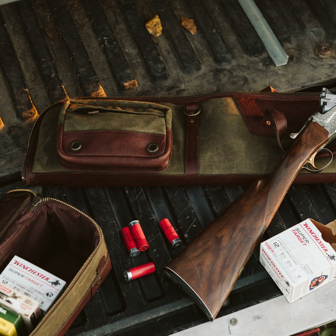 Overhead flat lay of an open waxed canvas shotgun case with a shotgun, loose shells, a box of cartridges and a leather bag
