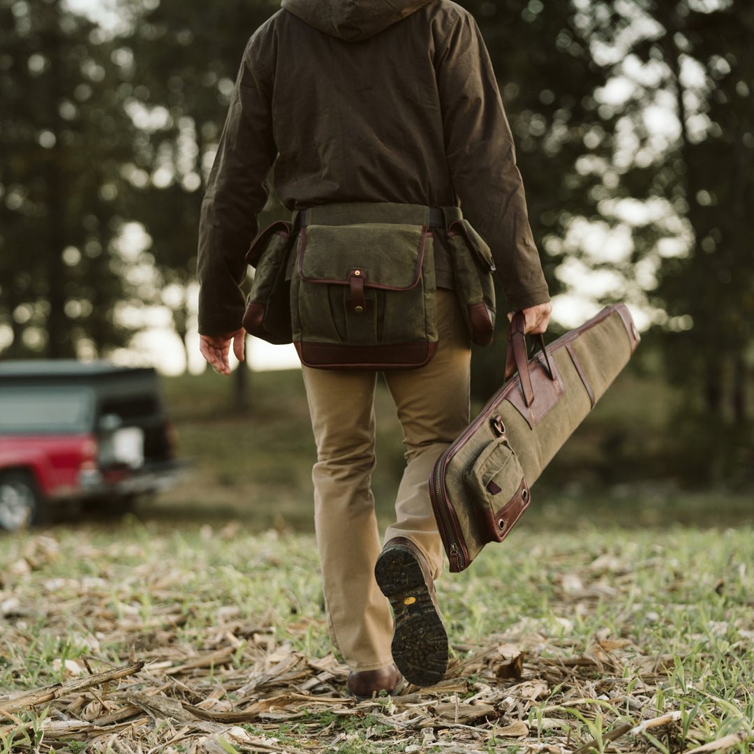Man walking away from a truck across a field carrying a waxed canvas shotgun case slung over his shoulder alongside a leather bag