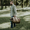 Outdoorsman with a rugged waxed canvas duffle bag in hand, set against a scenic woodland backdrop.