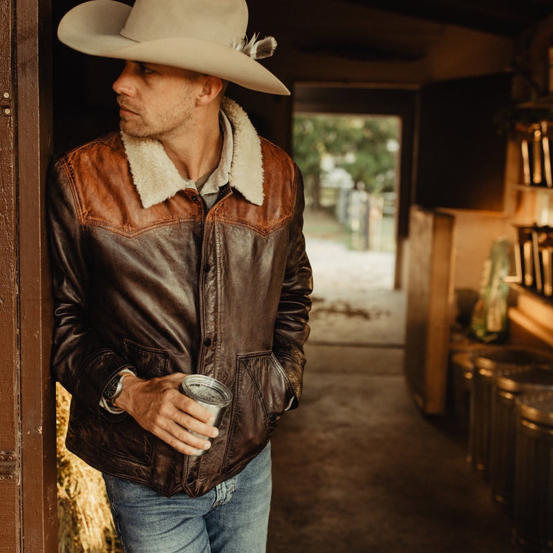 Man in a cowboy hat wearing a brown leather jacket with a shearling collar, standing in a rustic barn.